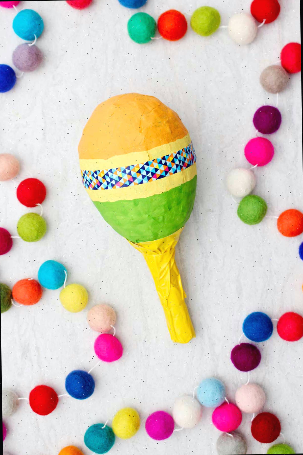 Maraca laranja, amarela e verde com cabo amarelo e fita washi colorida com pompons coloridos ao lado.