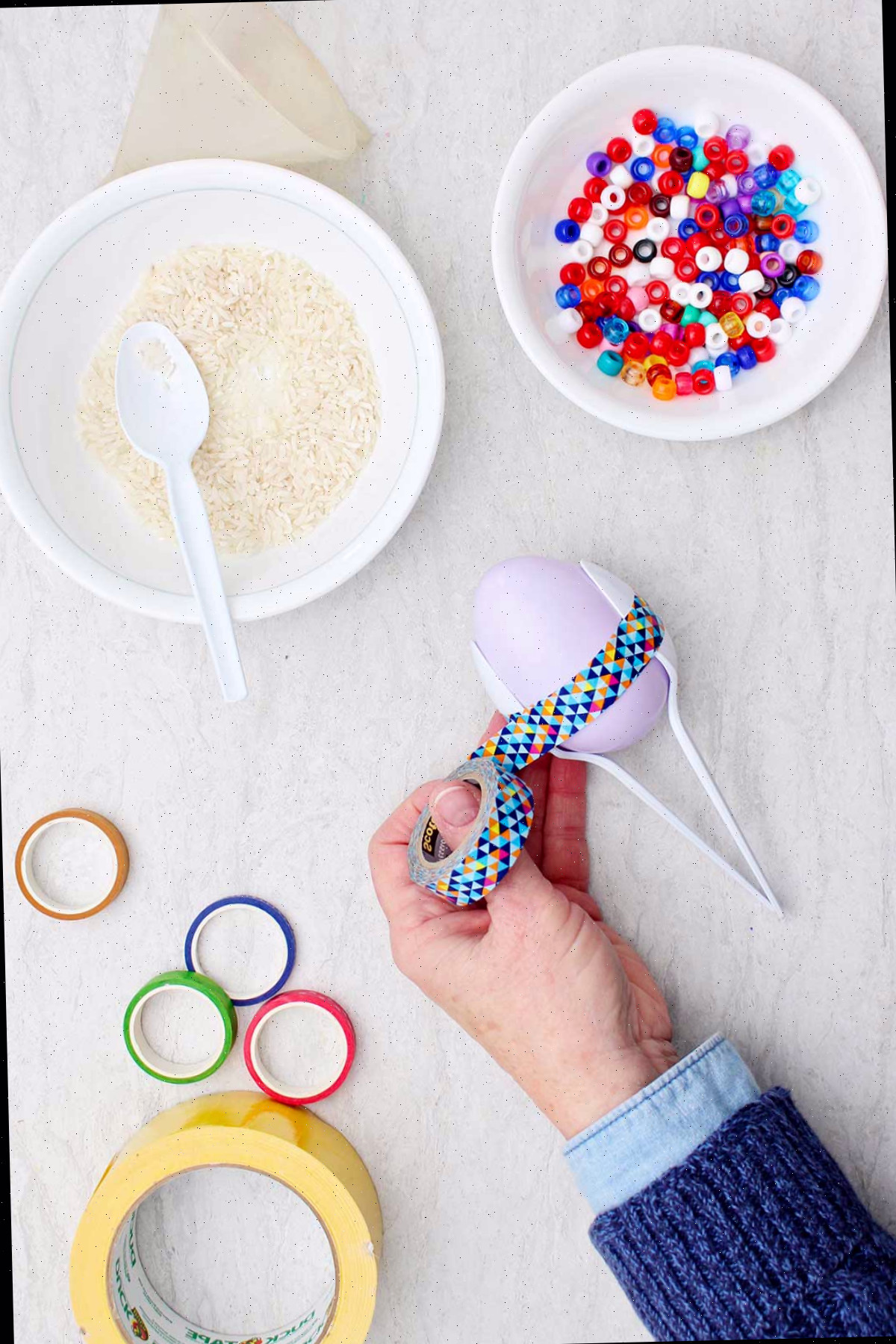 Indivíduo segurando duas colheres de plástico ao redor de um ovo de plástico com fita washi colorida.