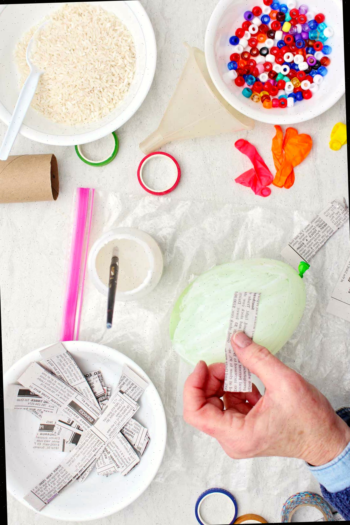 Pessoa colocando tiras de papel de jornal em um balão pintado com mod podge enquanto outros materiais estão ao redor.