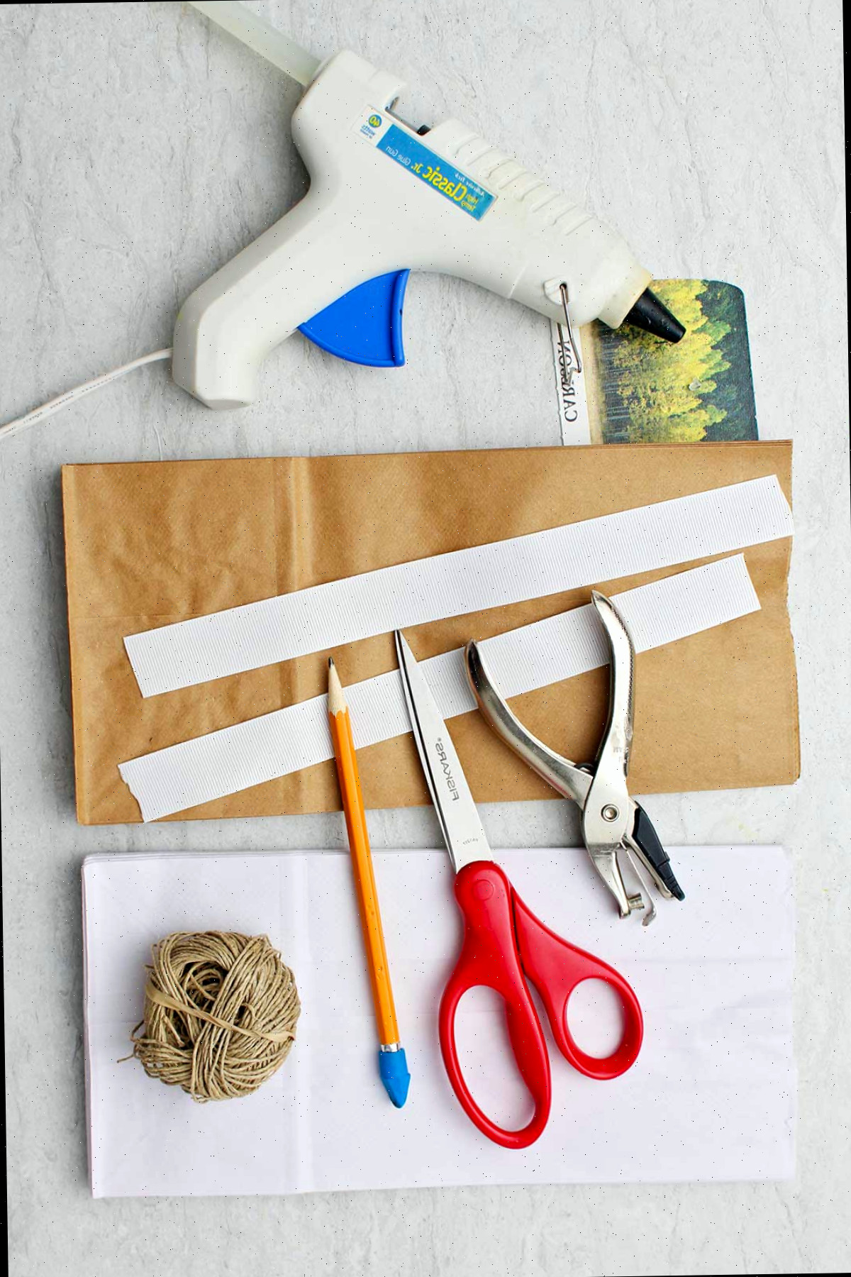 Materiais necessários para criar flocos de neve de papel. Fita adesiva, cola quente, perfurador, tesoura, lápis, barbante e sacos de papel.