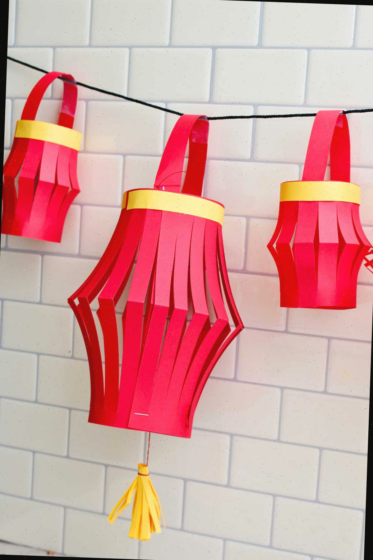 Três lanternas de papel concluídas penduradas contra um fundo de azulejos de metrô.