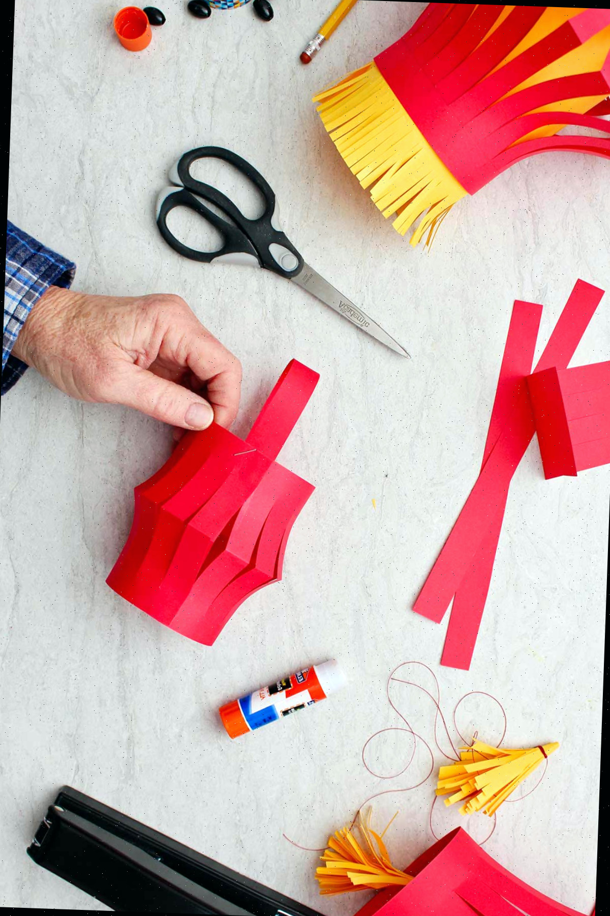 Mão segurando o corpo da lanterna de papel com uma tira de papel curvada sobre o topo, com outros suprimentos ao redor.