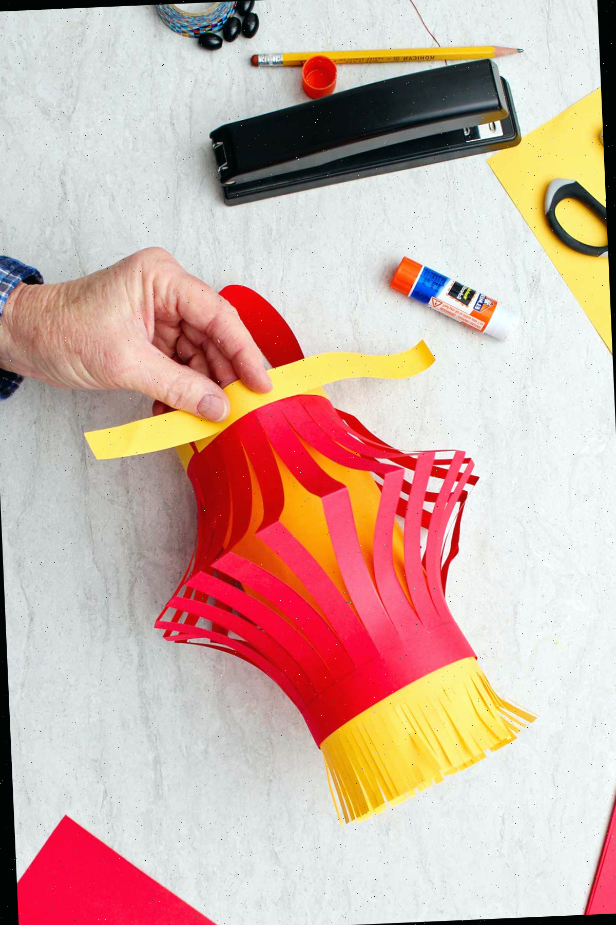 Mão colando uma tira de papel amarelo no topo da lanterna de papel para detalhes da decoração.