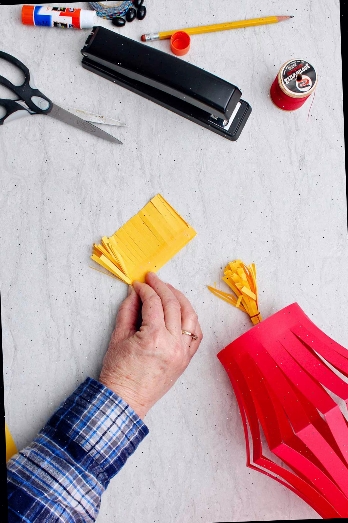 Mão dobrando a franja realizada com papel amarelo cortado, com suprimentos ao redor.
