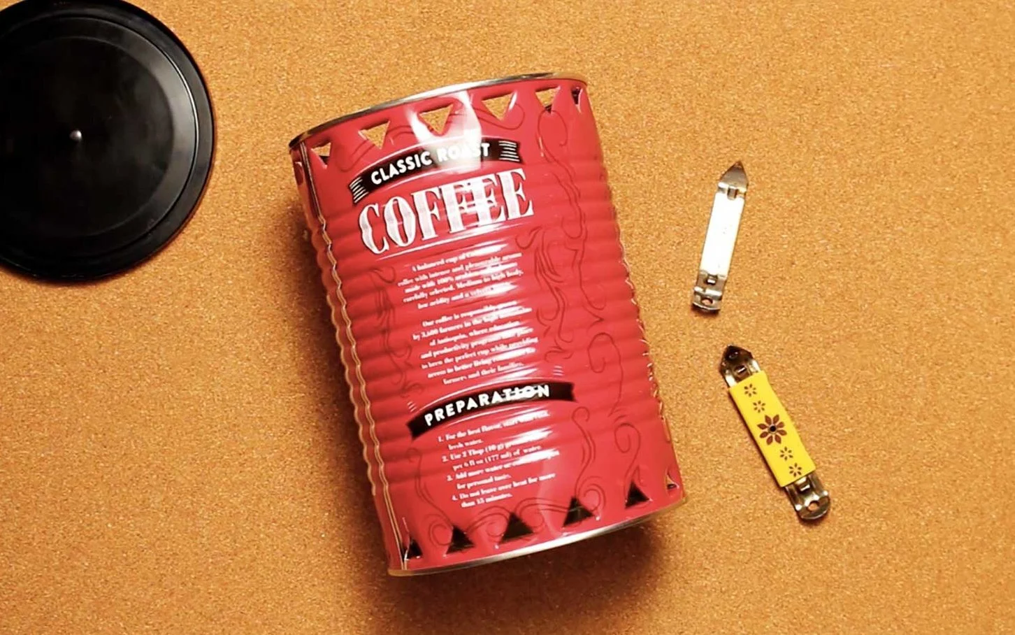 Uma lata de café, tampinha e dois pequenos abridores de lata repousando sobre um fundo cor de cortiça.