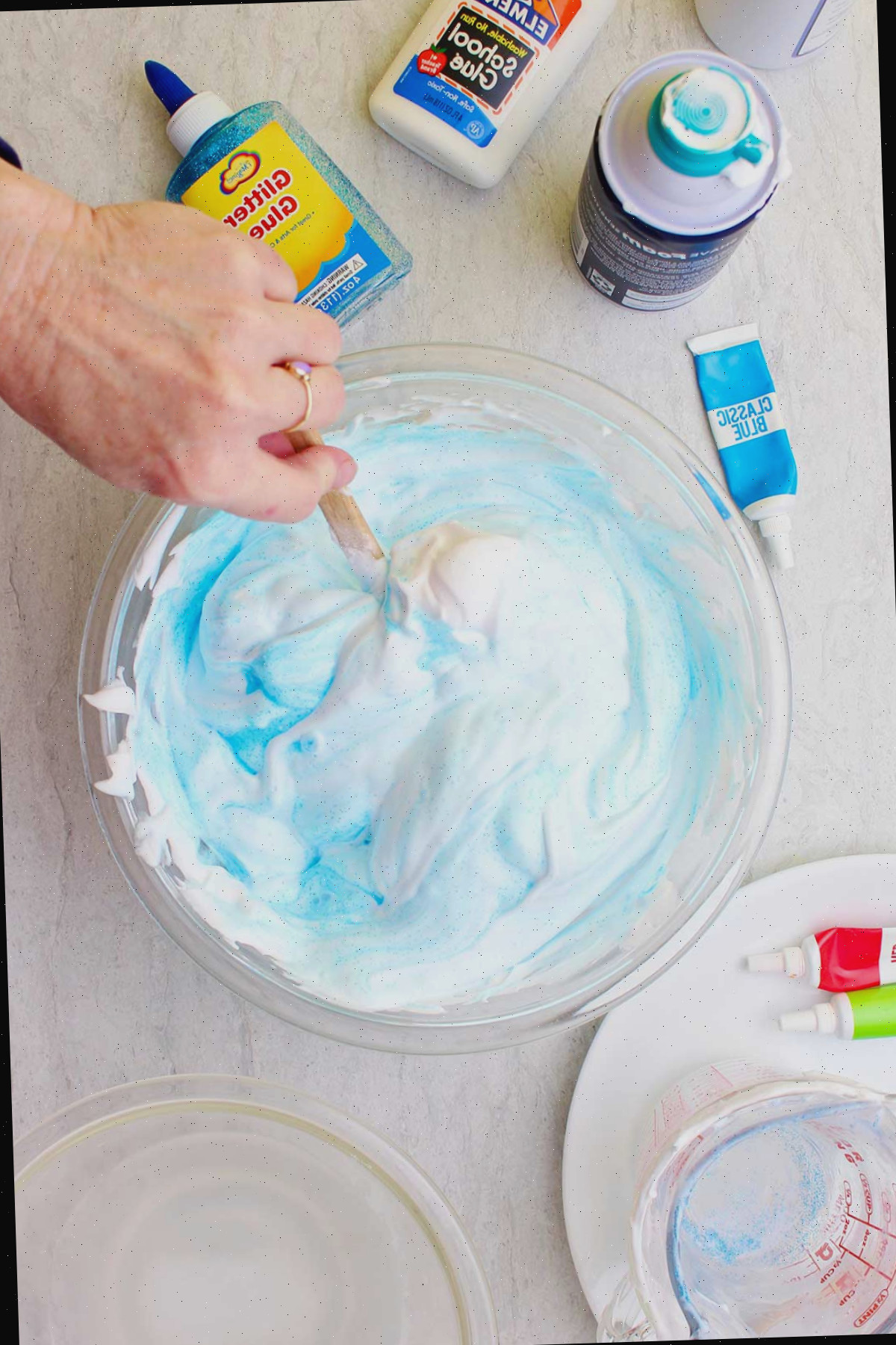 Mão misturando o corante em um lote de slime fofo azul claro em uma tigela de vidro com suprimentos espalhados ao redor.
