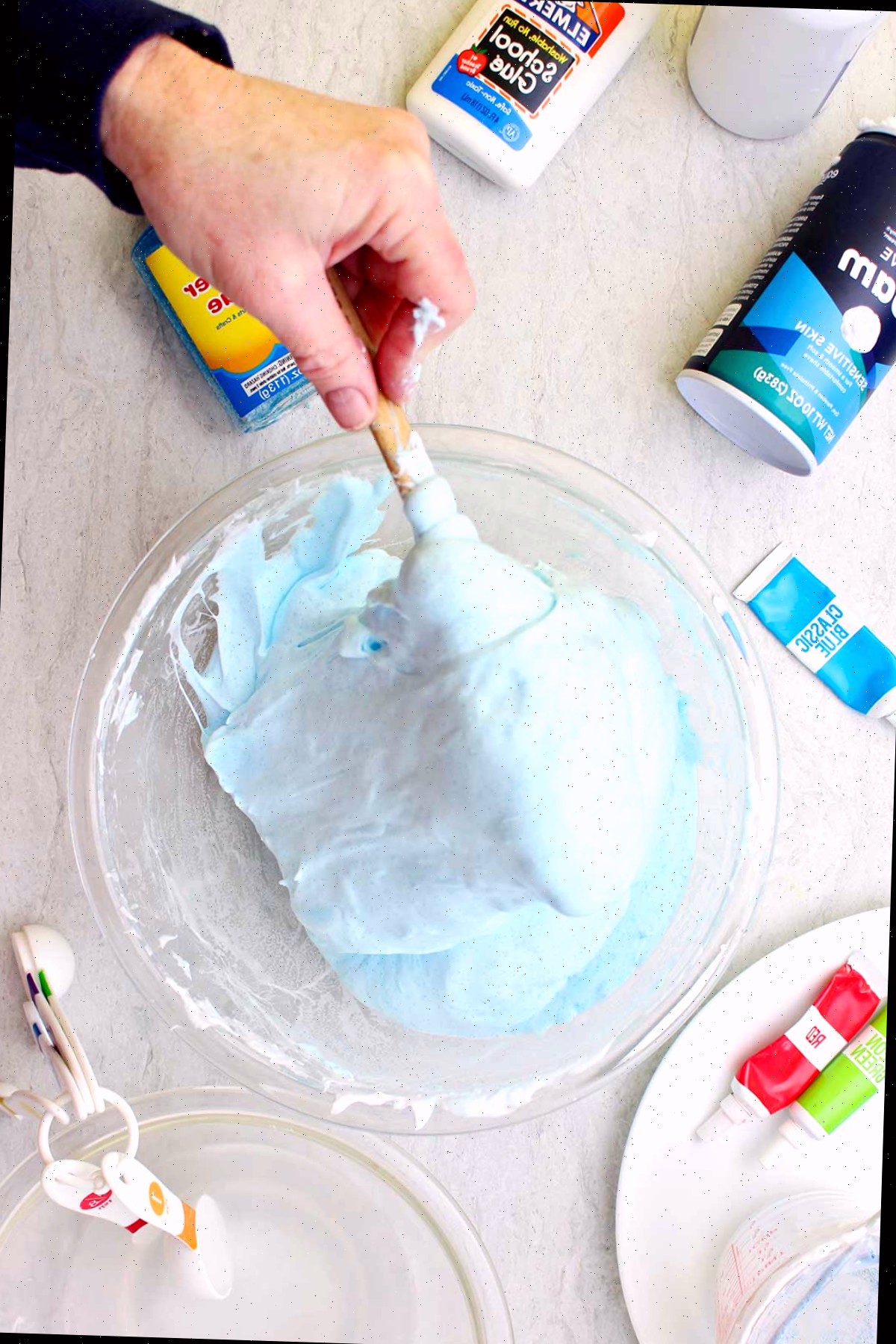 Mão mexendo um lote de slime fofo azul claro em uma tigela de vidro com suprimentos espalhados ao redor.