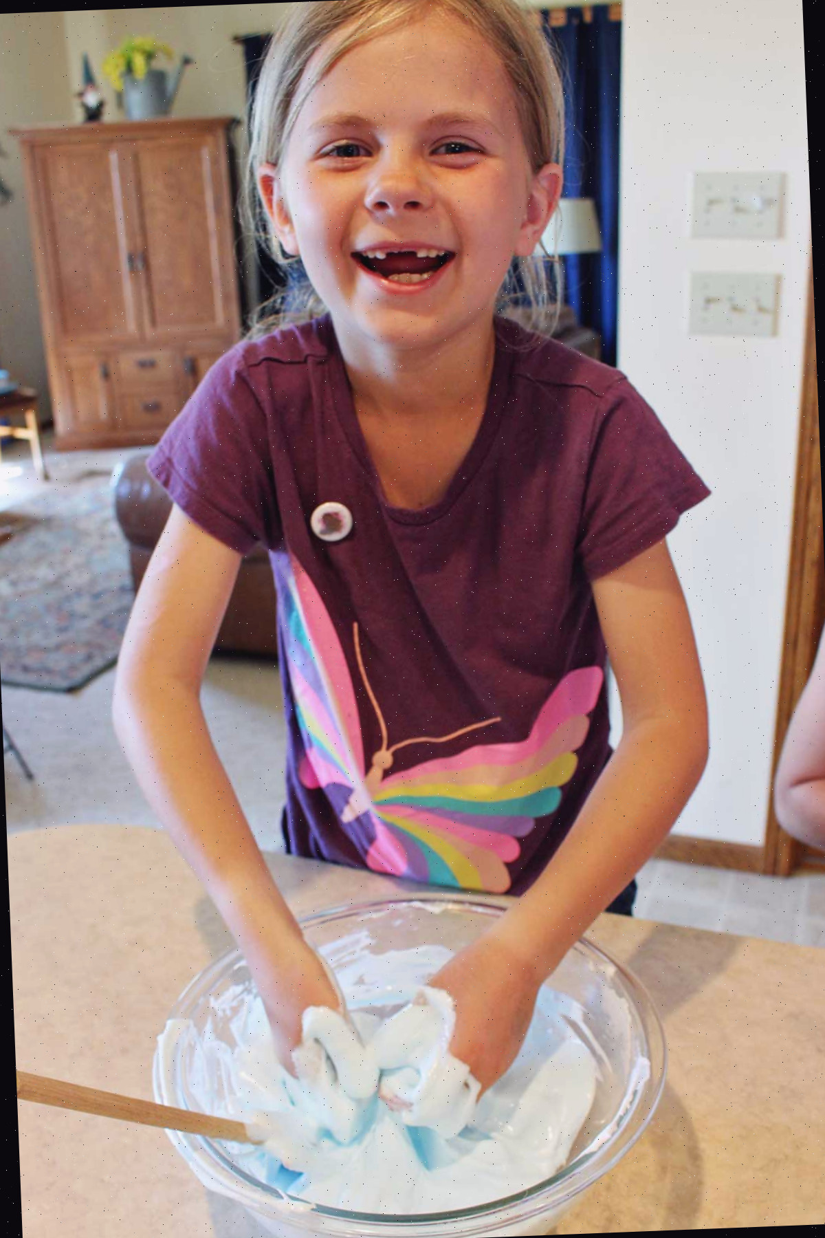 Menina jovem usando camisa de borboleta vinho sorrindo para a câmera brincando com um lote de slime fofo em uma tigela de vidro.