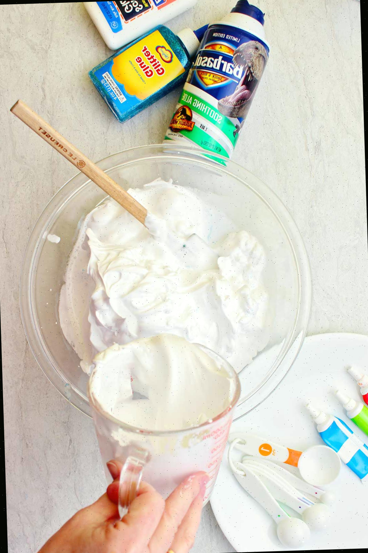 Mão despejando um componente em uma tigela de creme de barbear em uma tigela de vidro com suprimentos para slime fofo espalhados ao redor.