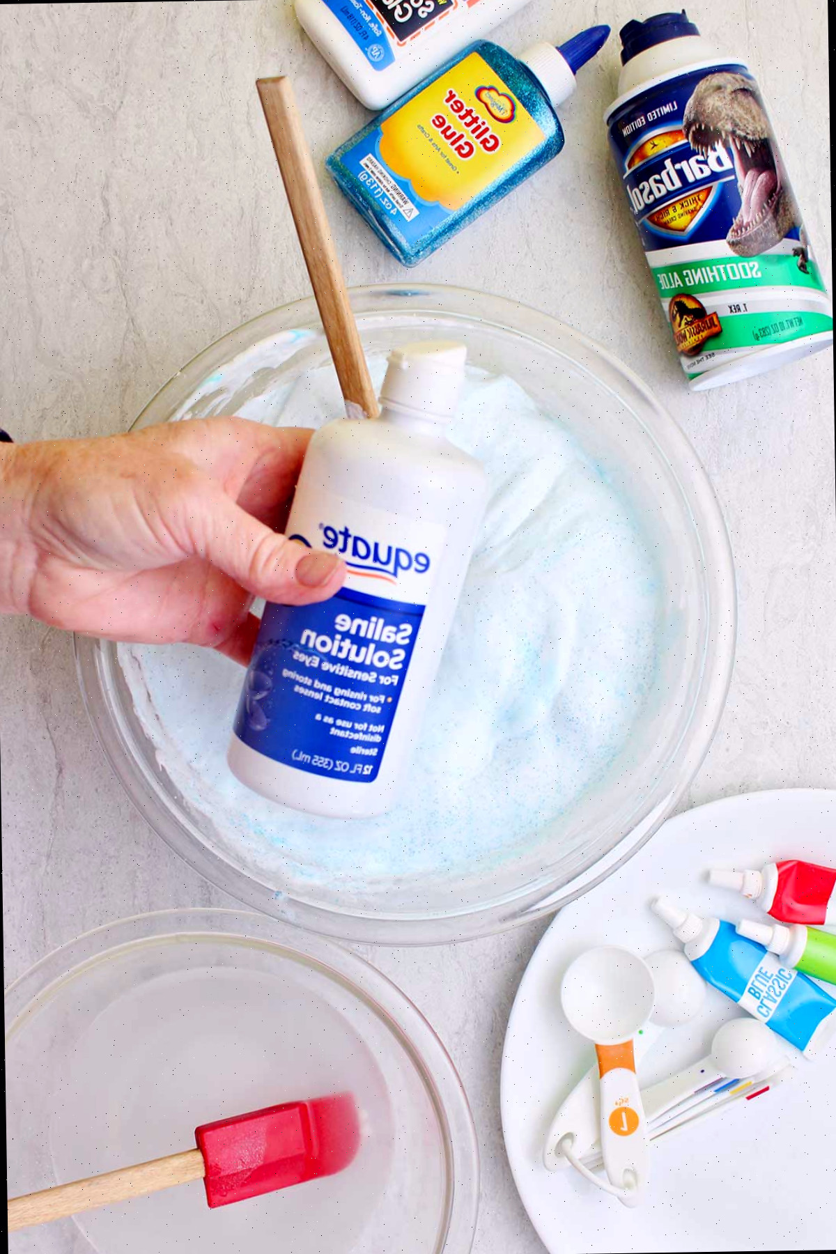 Mão segurando uma garrafa de solução salina acima de uma porção de slime fofo em uma tigela de vidro com suprimentos espalhados ao redor.