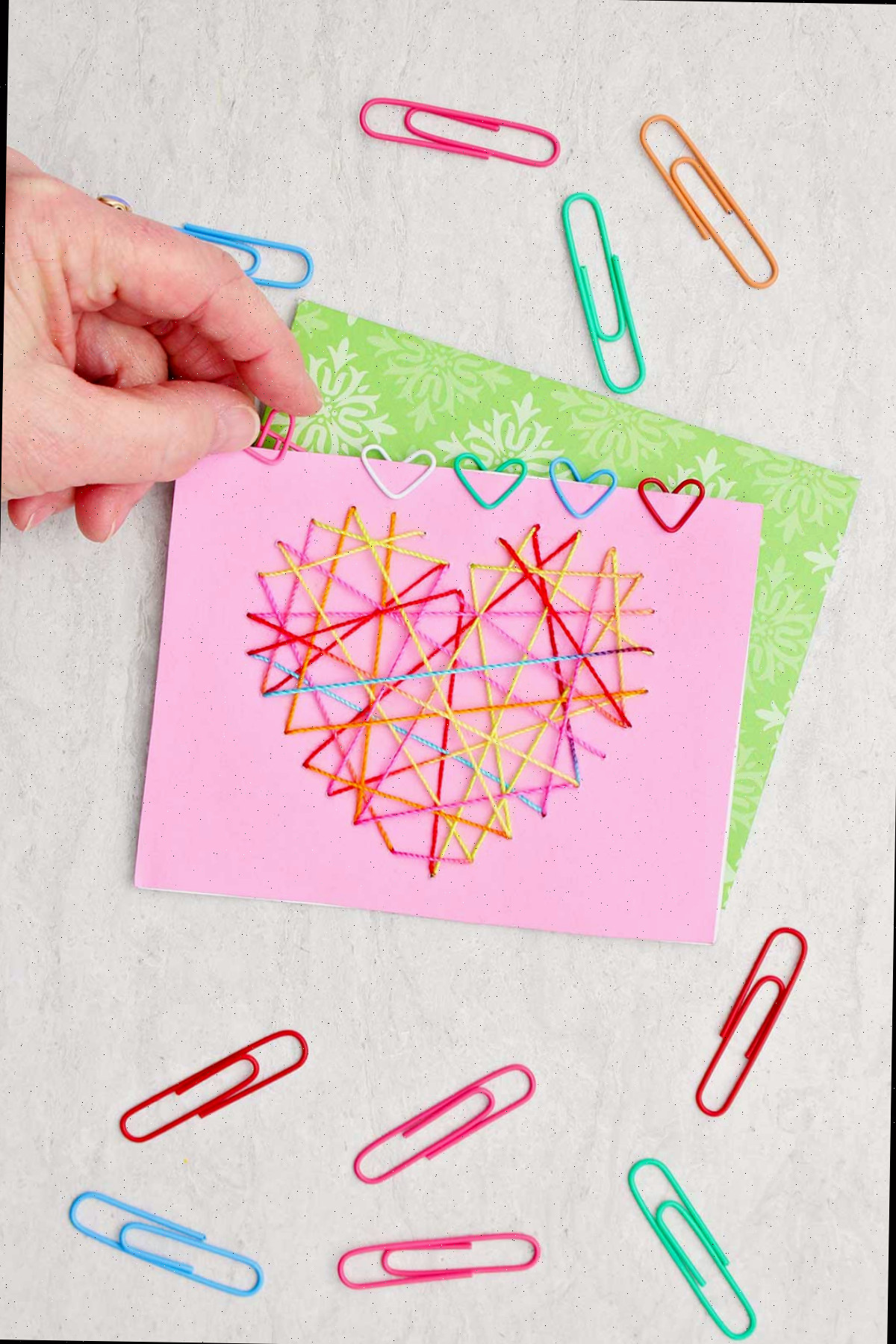 Indivíduo posicionando um clipe de papel em forma de coração rosa sobre um cartão de Dia dos Namorados com outros clipes de papel ao redor.