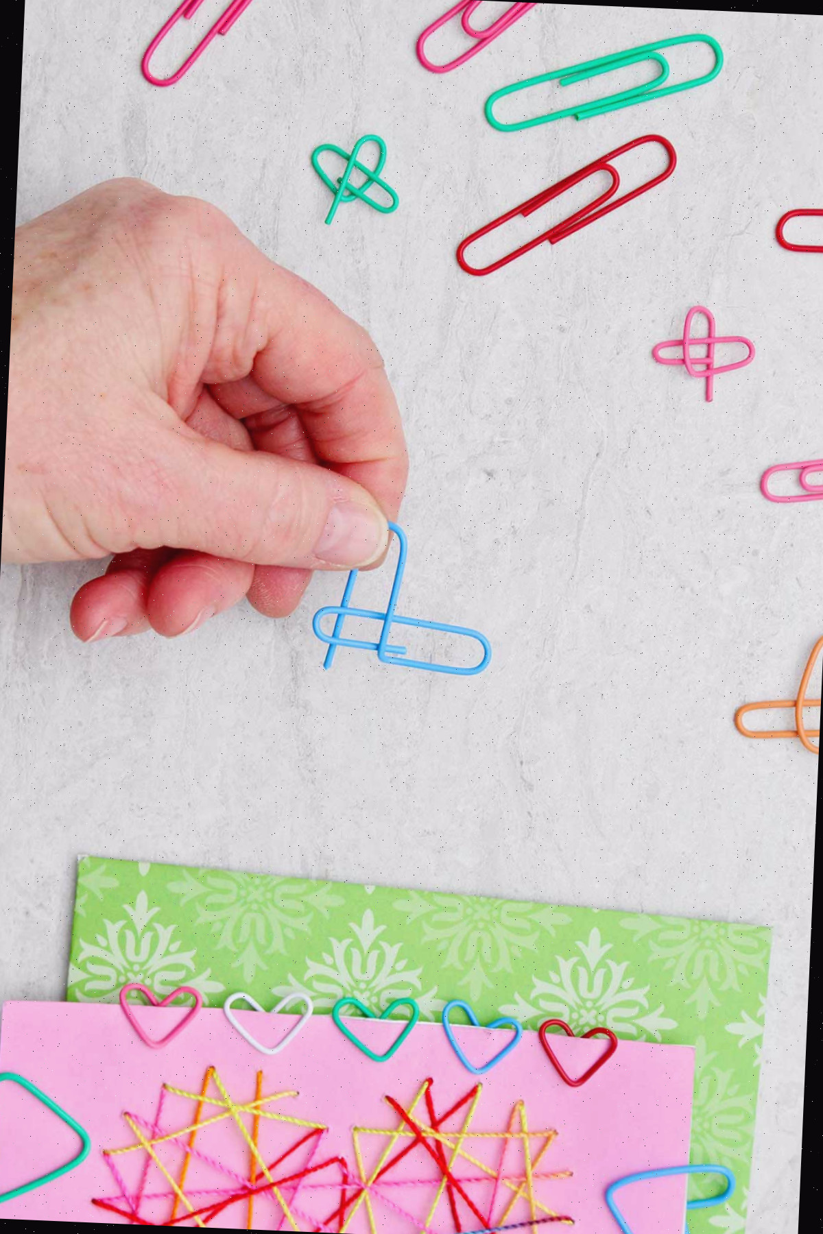 Pessoa demonstrando como dobrar um clipe de papel azul para tornar-se um coração, com outros clipes de papel coloridos e um cartão de Dia dos Namorados próximos.