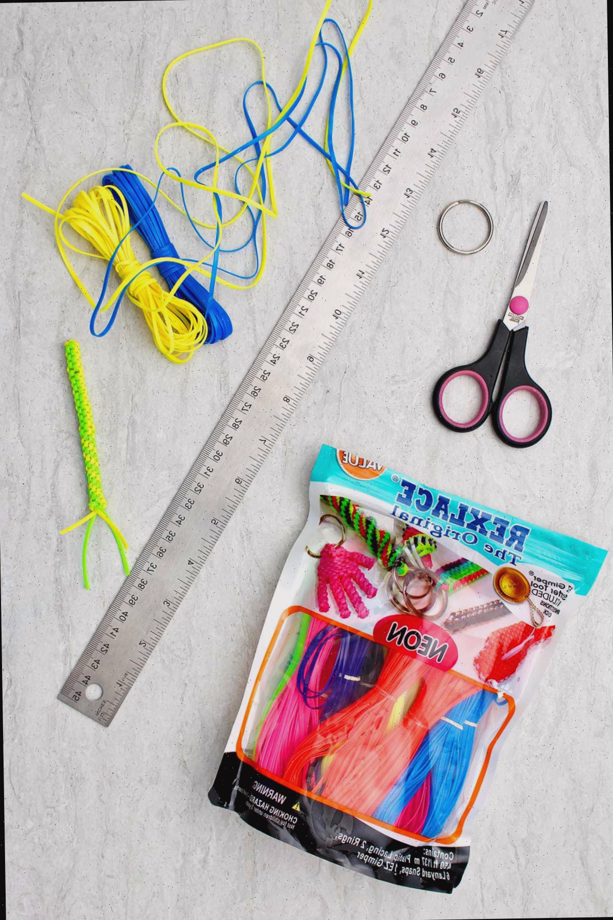Materiais necessários para confeccionar um Chaveiro Boondoggle. Rexlace neon, tesoura, uma régua de metal e um chaveiro.