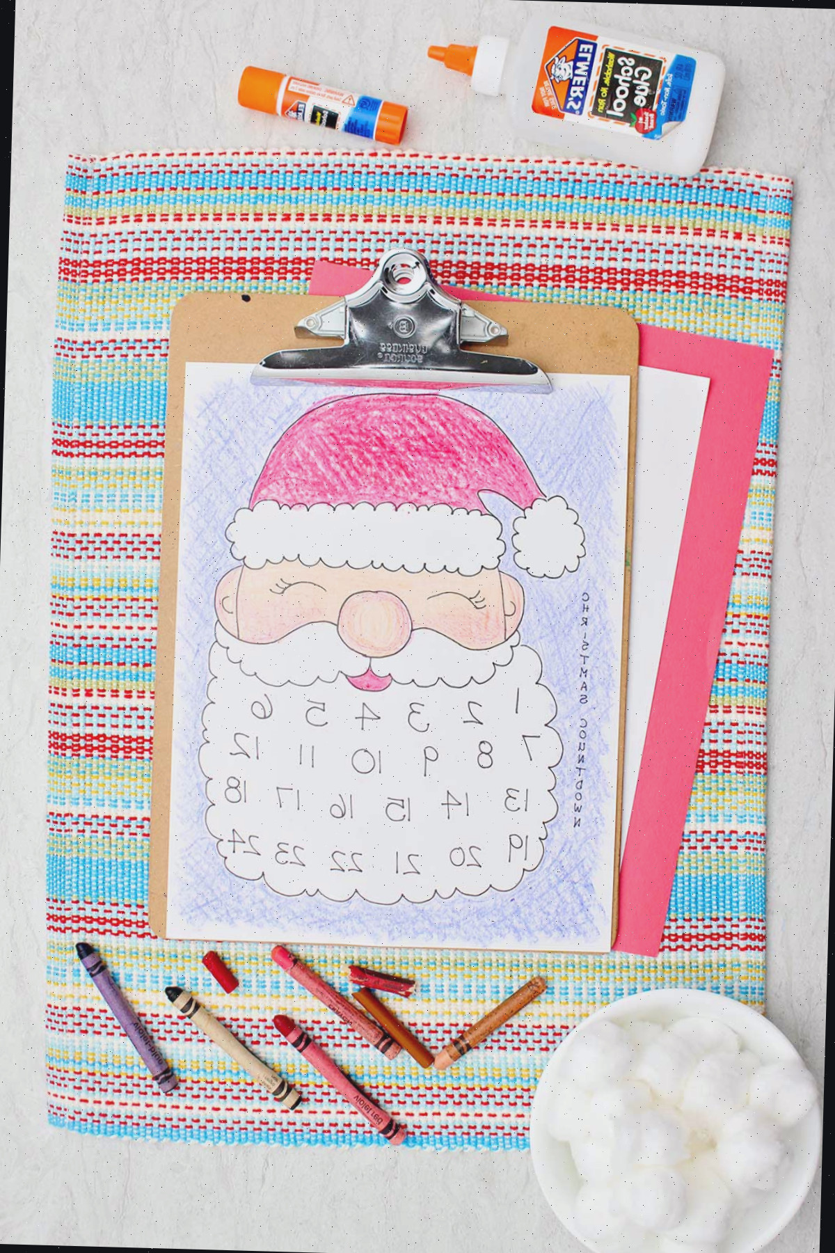 Um calendário vibrante do Papai Noel impresso em uma prancheta sobre um jogo americano com cola, giz de cera e bolinhas de algodão ao lado.