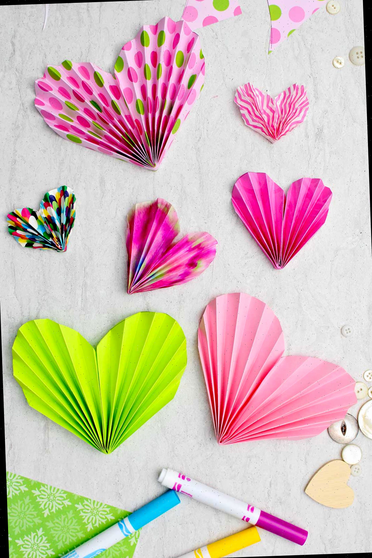 Acordeões de papel rosa e verde usados para ornamentar cartões e valentins, com algumas canetas e botões dispersos nas imediações.