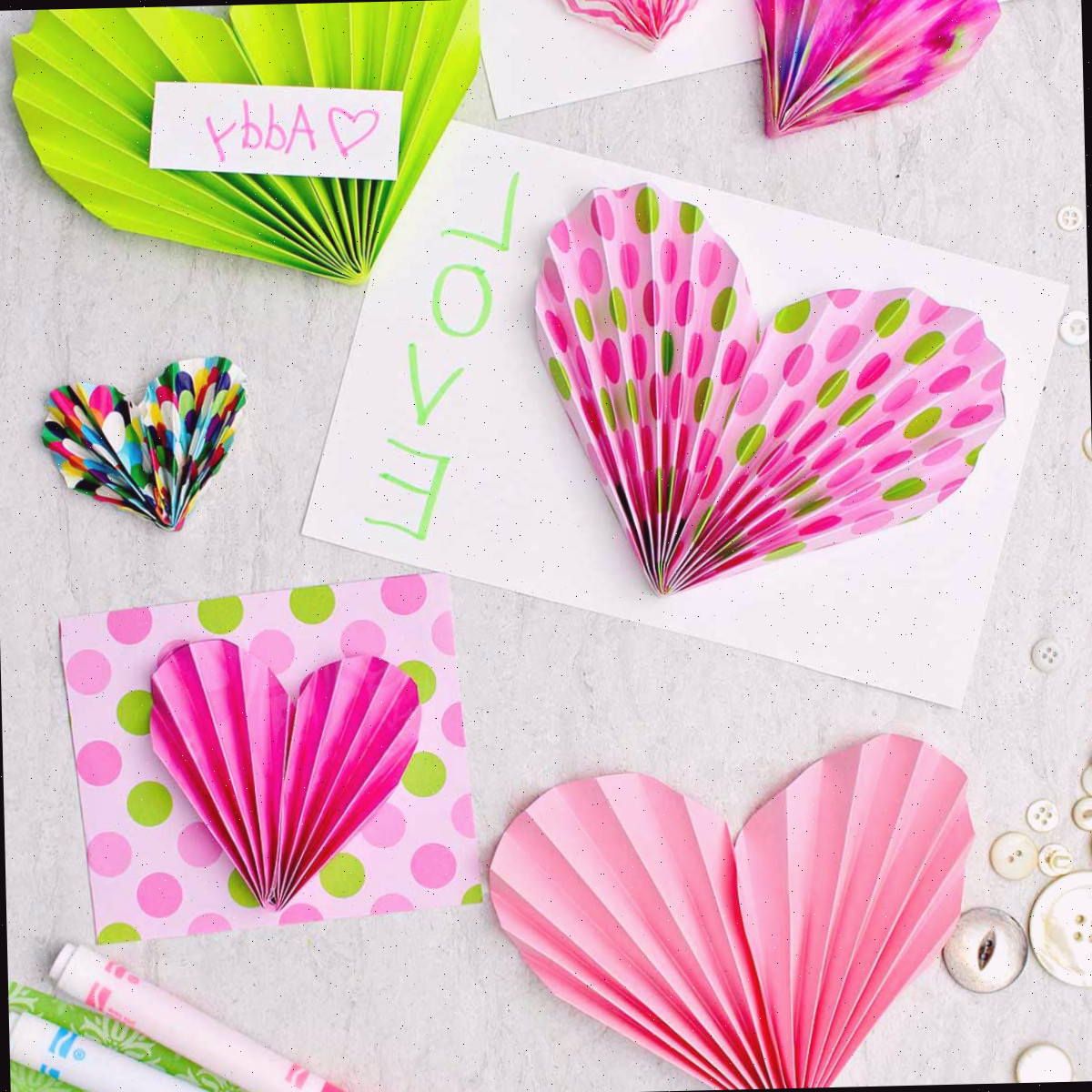 Acordeões de papel rosa e verde usados para ornamentar cartões e valentins, com algumas canetas e botões dispersos nas imediações.