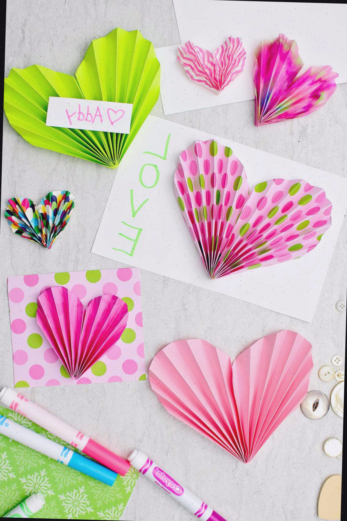 Corações de papel em tons de rosa e verde usados para enfeitar cartões e valentins com alguns marcadores e botões espalhados nas imediações.
