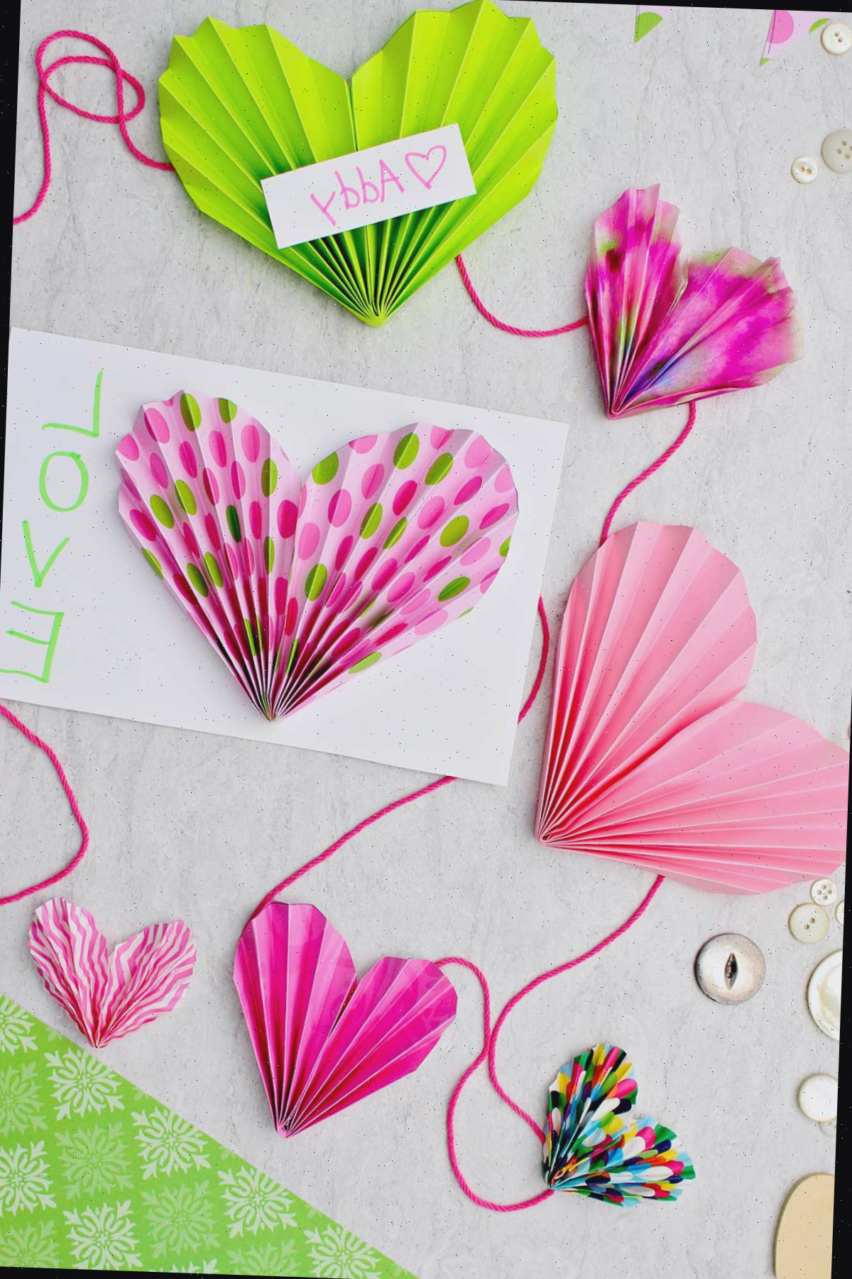 Corações de papel sanfonado rosa e verde finalizados. Alguns criados para cartões de Dia dos Namorados e outros presos a uma corda para formar uma guirlanda.