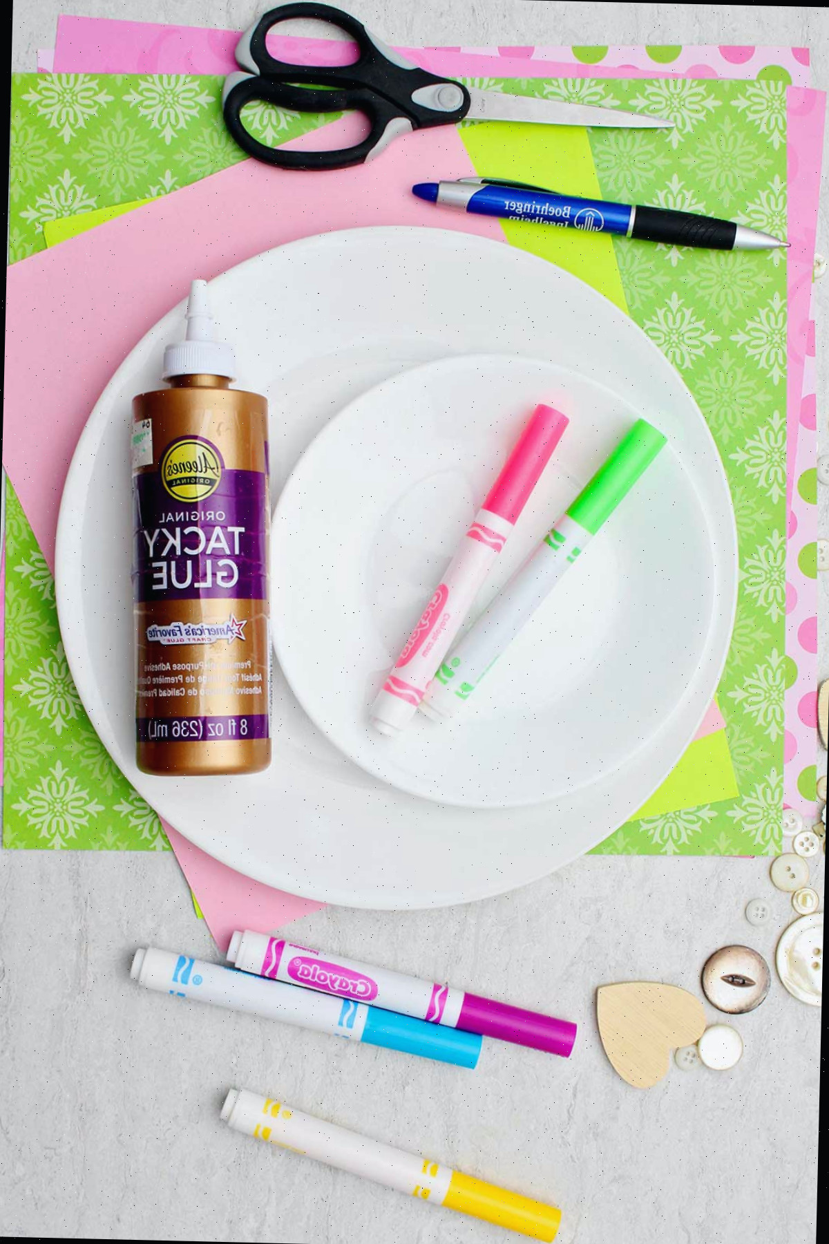 Cola, papel estampado verde e rosa, um prato, marcadores, tesoura e botões.