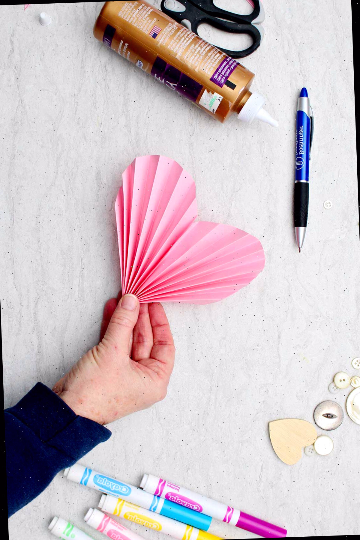 Indivíduo segurando um coração de papel em acordeão rosa com outros materiais ao redor.