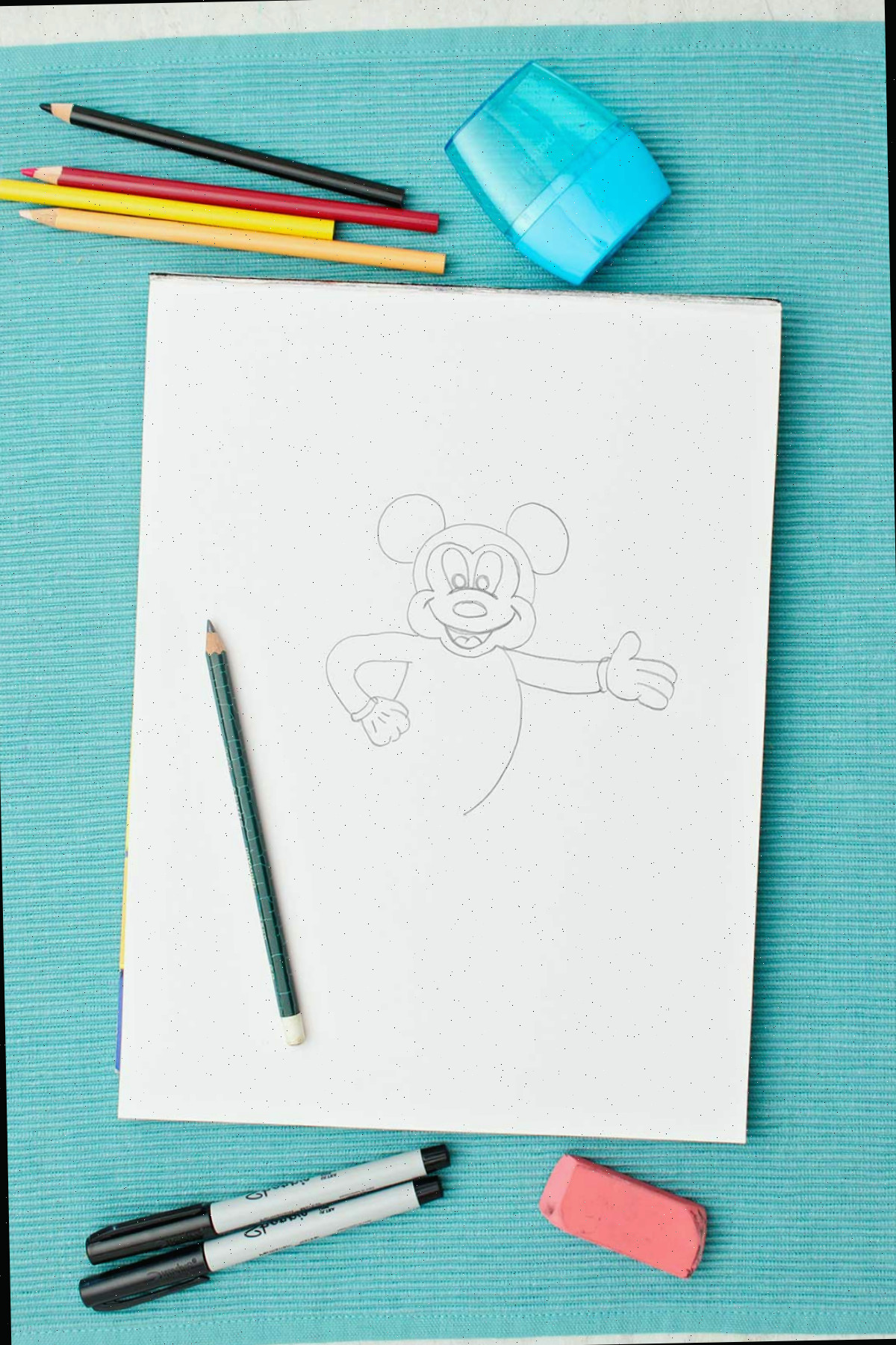 Um esboço a lápis semi-completo do Mickey Mouse e alguns lápis de cor e materiais ao lado.