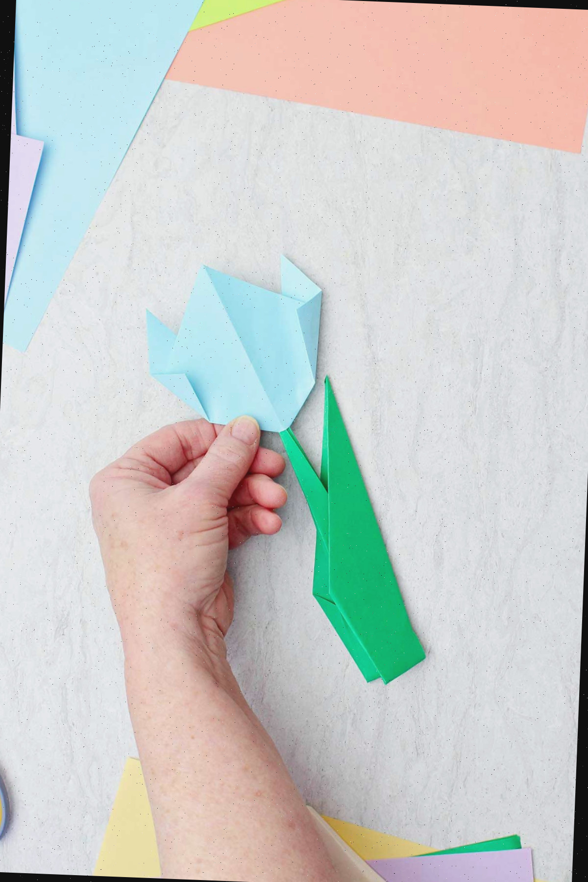 Uma pessoa segurando uma flor de origami azul clara, com outros papéis coloridos ao redor.