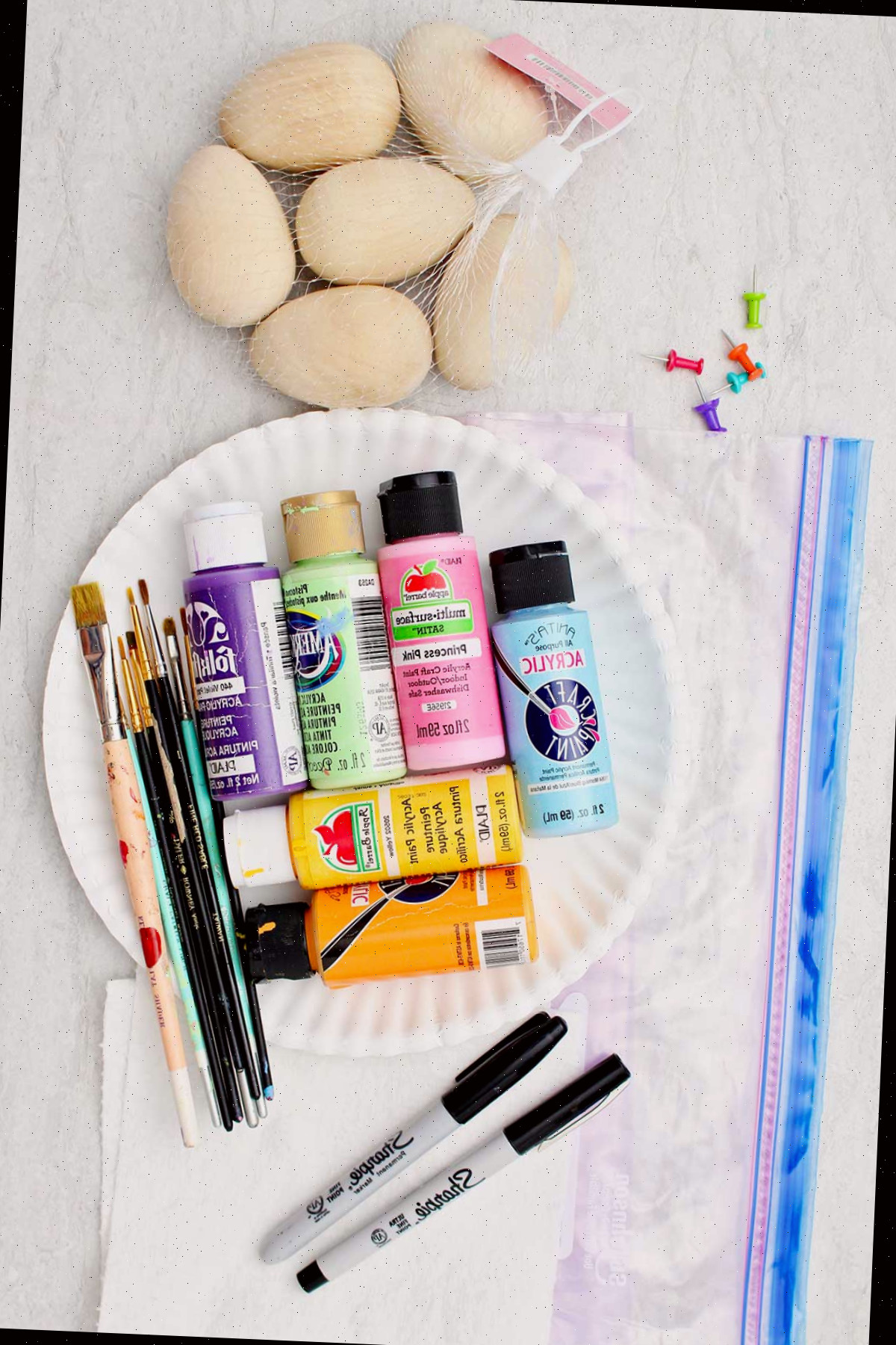 Uma bolsa contendo ovos de madeira, um saco plástico com zip-lock, tintas acrílicas coloridas, pincéis e marcadores sharpie.