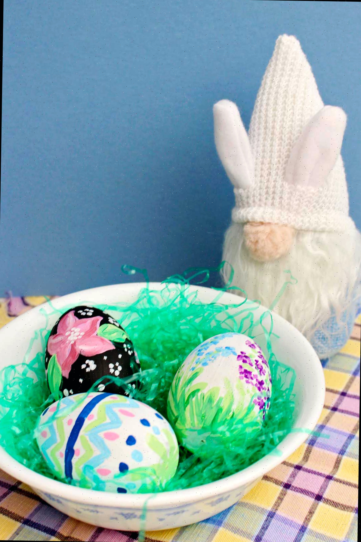Uma tigela com três ovos de madeira coloridos na grama de uma cesta de Páscoa repousando sobre um pano quadriculado pastel com um gnomo coelho próximo.