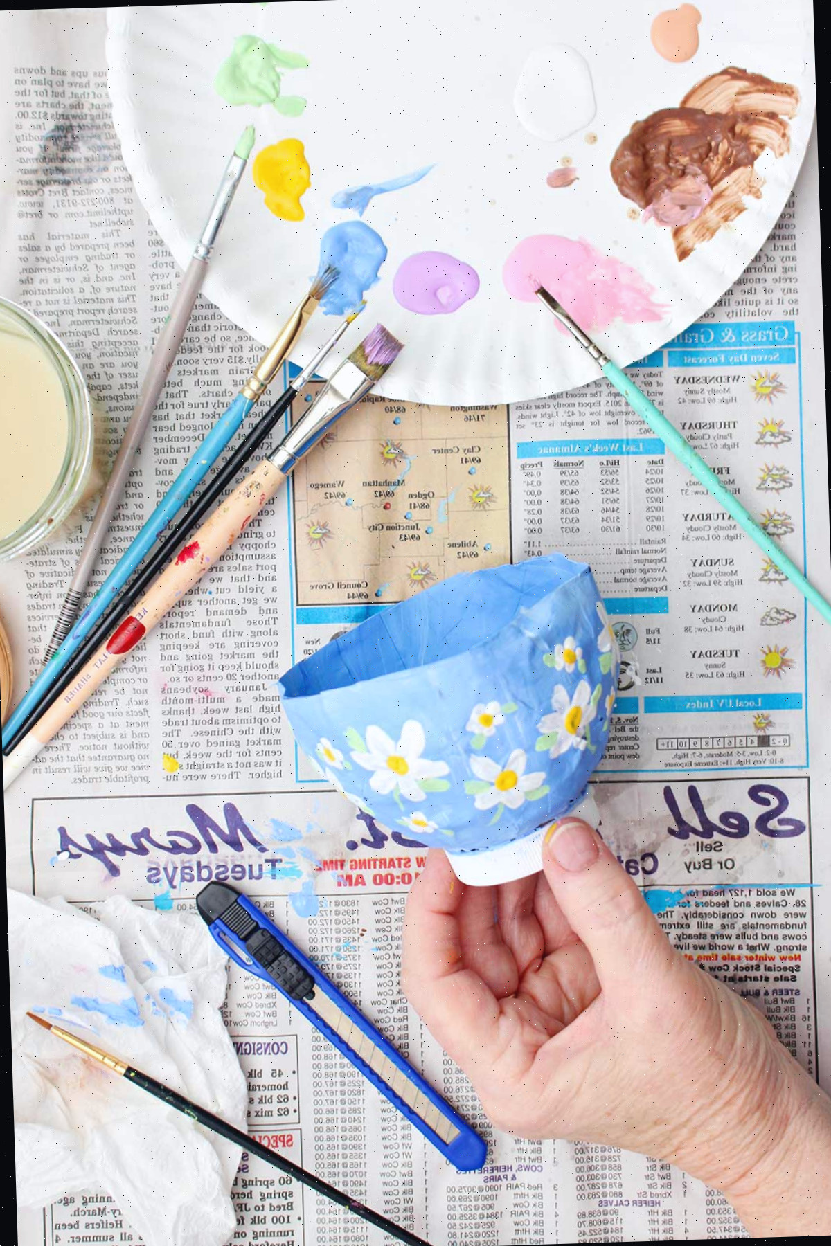 Uma pessoa segurando um projeto de papel machê pintado de fundo azul claro com flores brancas e amarelas.