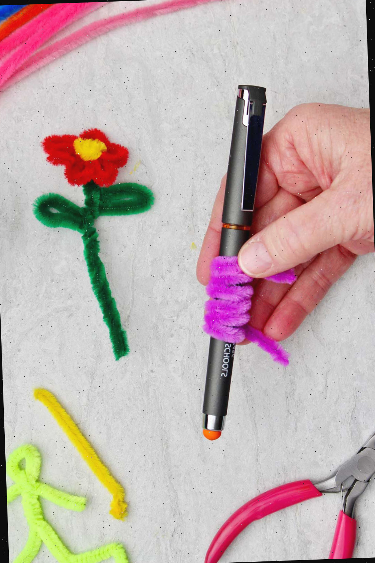Uma pessoa segurando uma caneta com um limpador de tubo roxo enrolado ao seu redor, com uma flor de limpador de tubo e outros limpadores de tubo ao fundo.