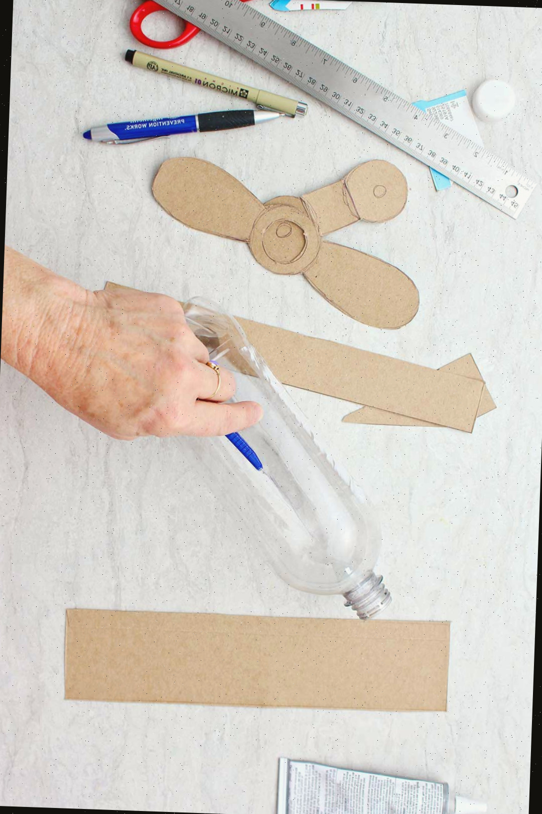 Mão usando um cortador para criar um corte na garrafa plástica vazia com asas de papelão e hélices próximas.