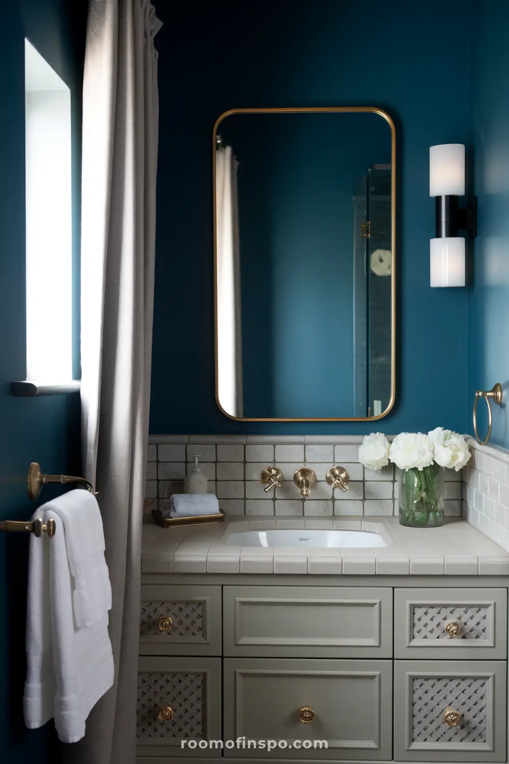 Um banheiro elegante com paredes azul-escuras, um amplo espelho retangular emoldurado em dourado, uma pia branca com acessórios dourados, uma bancada de azulejos brancos, um vaso com flores brancas e uma luminária com duas lâmpadas cilíndricas.