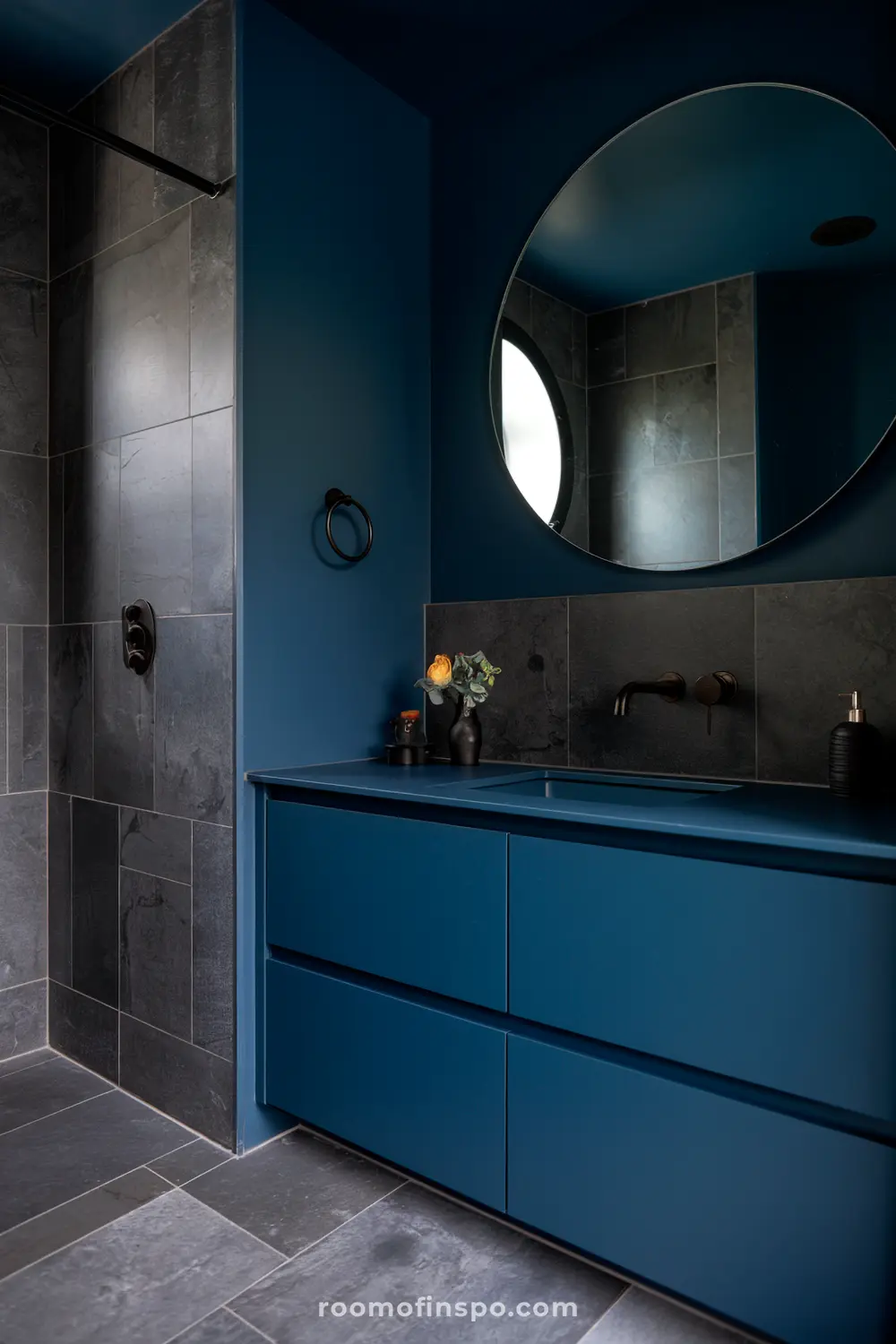 Um banheiro contemporâneo com armários azul escuro, um grande espelho arredondado, uma torneira preta e uma área de banho com azulejos cinza escuro.
