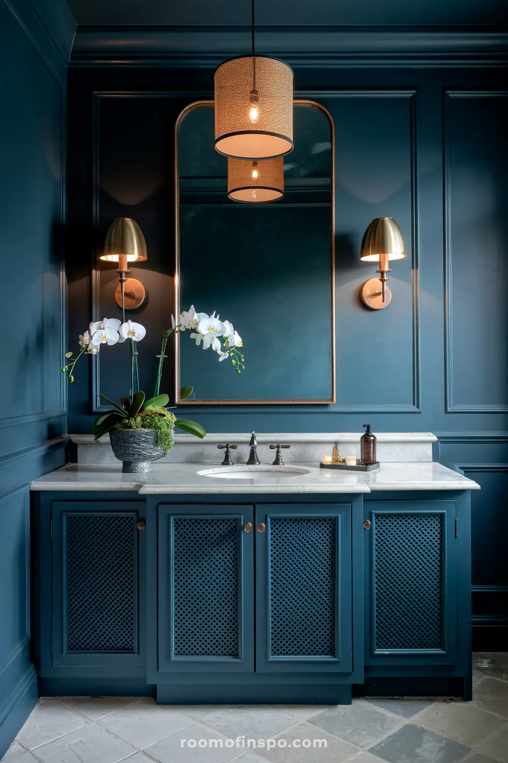 Uma vaidade de banheiro elegante com uma bancada em mármore, apresentando uma pia, uma orquídea em vaso, uma bandeja com produtos de higiene, rodeada por dois apliques de parede em bronze, tudo contra paredes cobertas em azul profundo com um grande espelho e uma luminária suspensa acima.
