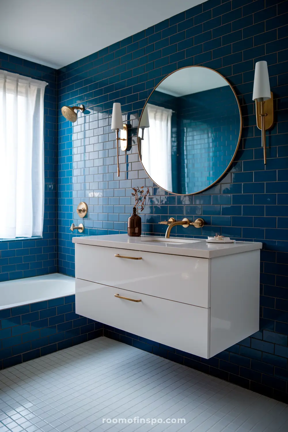 Um banheiro contemporâneo com paredes de azulejos azul em forma de ladrilho, um espelho redondo, uma vaidade flutuante branca com acessórios dourados, uma torneira de parede, e dois apliques de parede, apresentando uma janela com cortinas brancas e uma banheira.