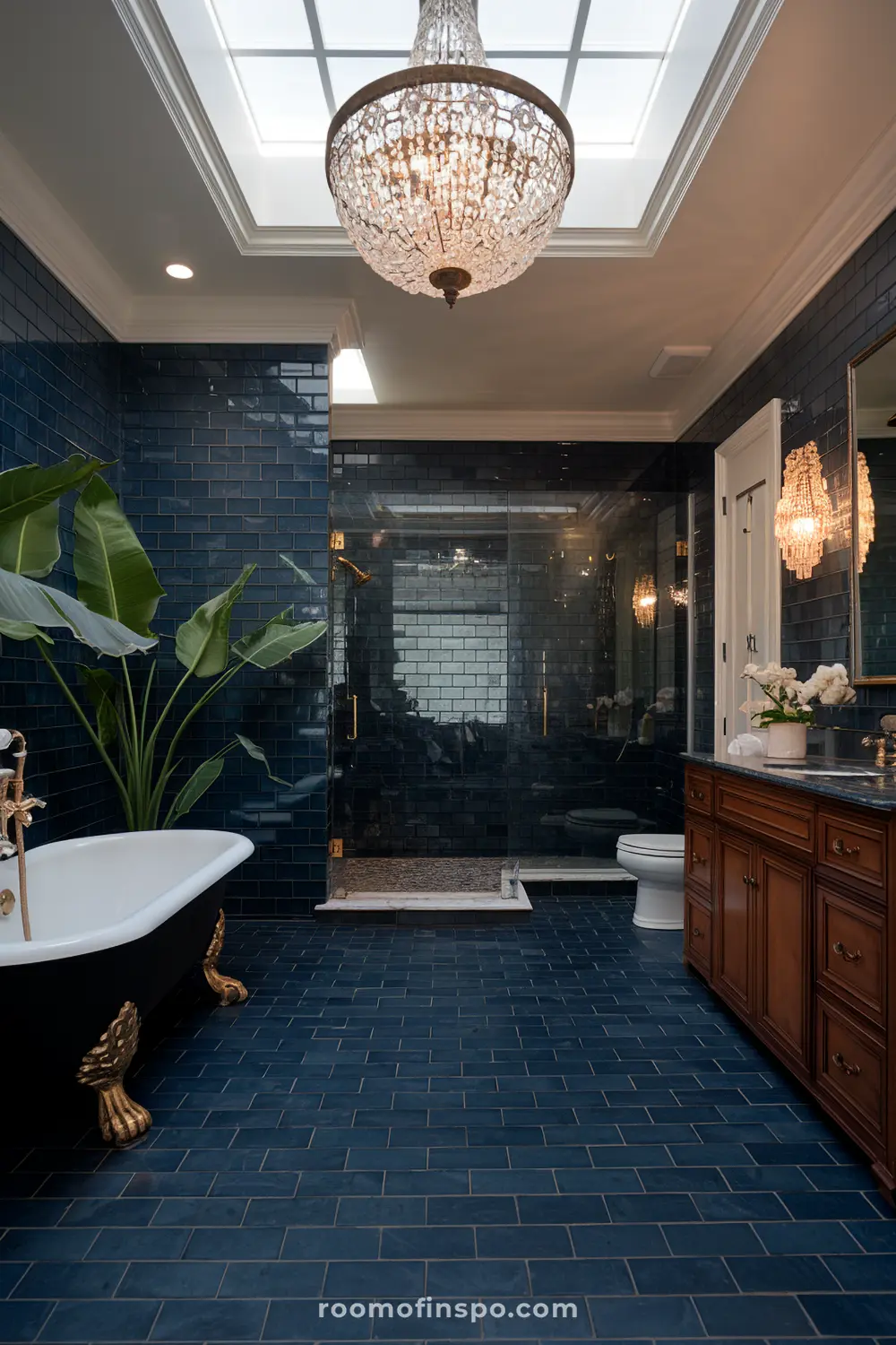 Um banheiro sofisticado com ladrilhos escuros em formato de azulejo, uma banheira isolada com pés dourados, uma planta grande, uma vaidade de madeira com uma superfície de mármore, um vaso sanitário, um chuveiro envidraçado, e um candelabro suspenso em uma claraboia.