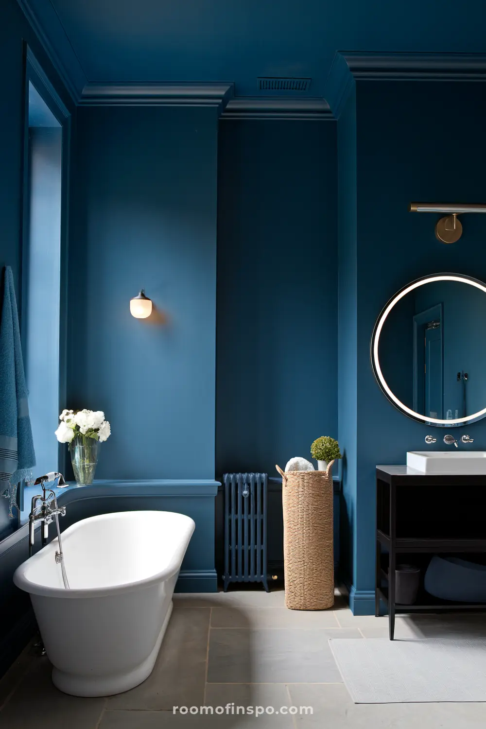 Um banheiro contemporâneo com paredes azul-escuras que exibe uma banheira branca independente, uma luminária de parede montada, um vaso com flores brancas, uma cesta de vime, um espelho redondo sobre uma vaidade preta com uma pia branca, e um radiador.