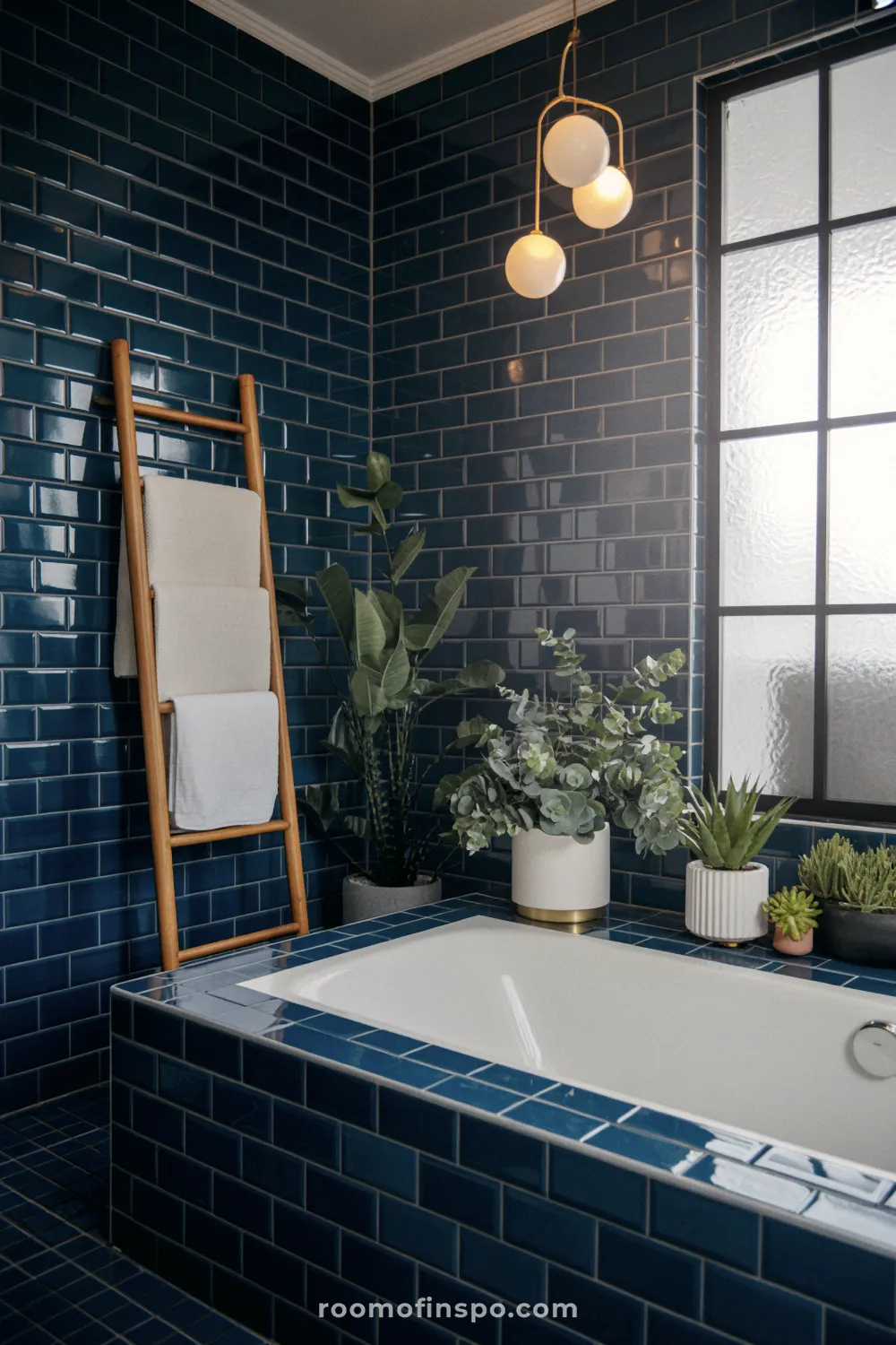 Banheiro em azul escuro com azulejos brilhantes em formato de ladrilho, uma banheira embutida, uma escada de madeira para toalhas e vegetação sob uma janela escura.
