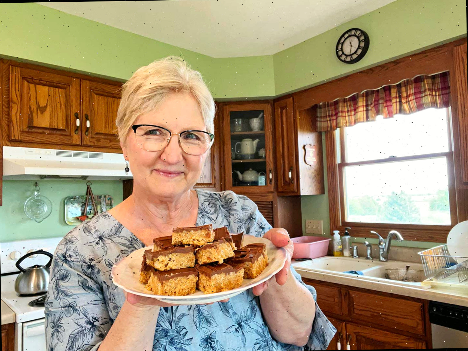 Nana sorrindo e segurando um prato de scotcharoos em sua cozinha.