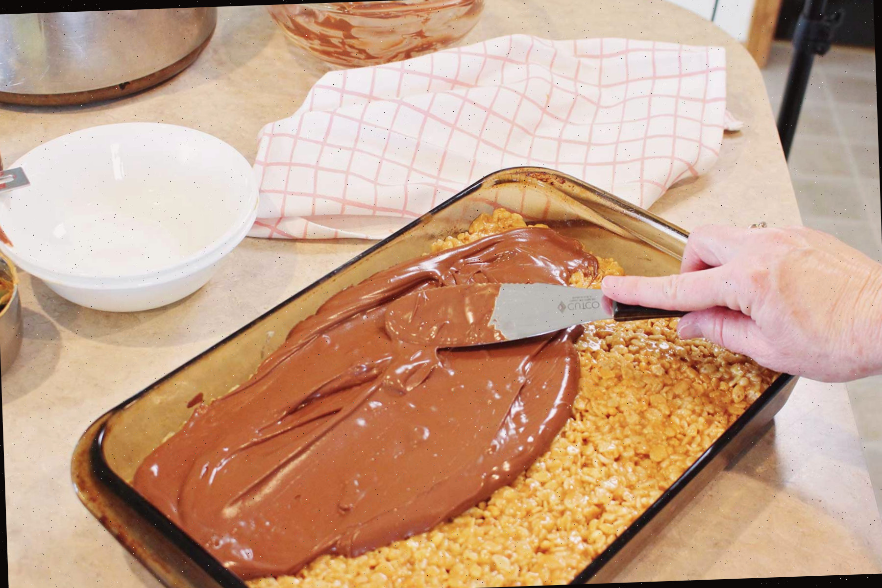 Uma pessoa utilizando uma espátula de metal para espalhar chocolate derretido sobre os scotcharoos.
