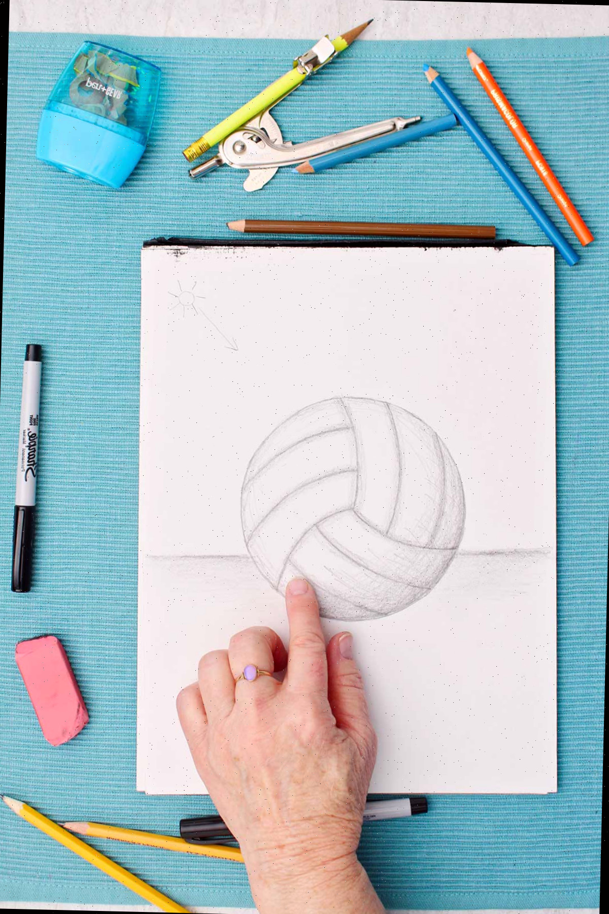 Desenho de uma bola de vôlei a lápis com uma pessoa sombreadando com o dedo e materiais ao lado em uma mesa azul.