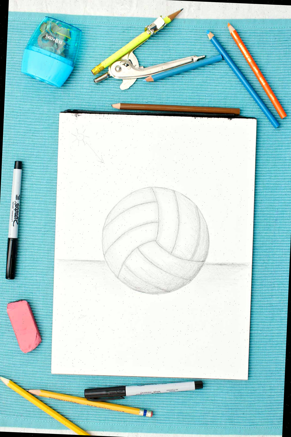 Desenho de uma bola de vôlei a lápis com sombreamento e materiais ao lado em uma mesa azul.