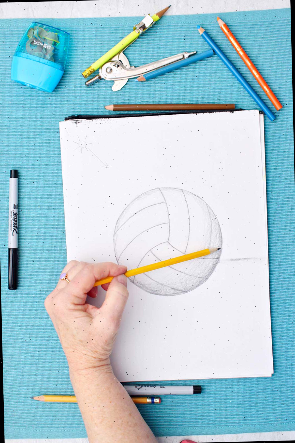 Desenho de uma bola de vôlei esboçada com lápis, enquanto uma pessoa faz sombreamento com o dedo e materiais ao redor em uma mesa azul.
