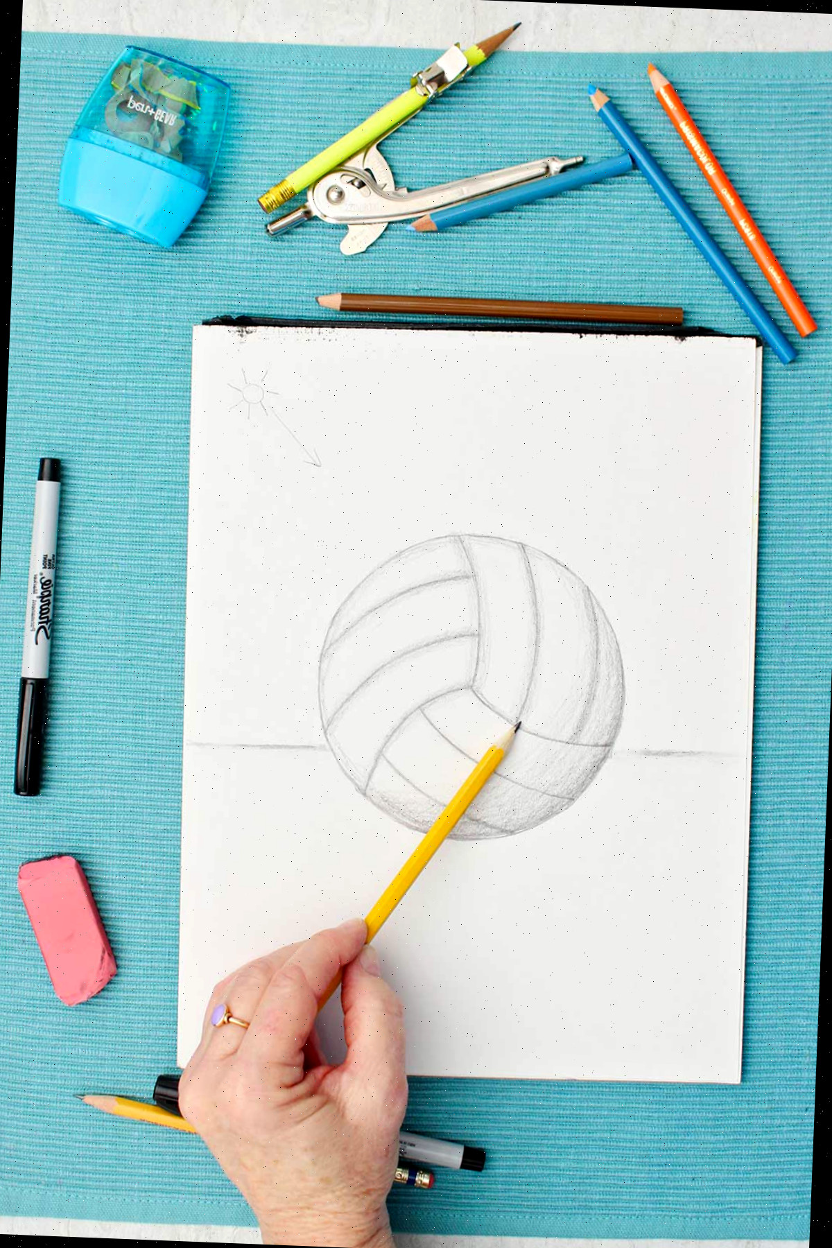Desenho de uma bola de vôlei esboçada com lápis, enquanto uma pessoa faz sombreamento usando lápis e materiais ao redor em uma mesa azul.