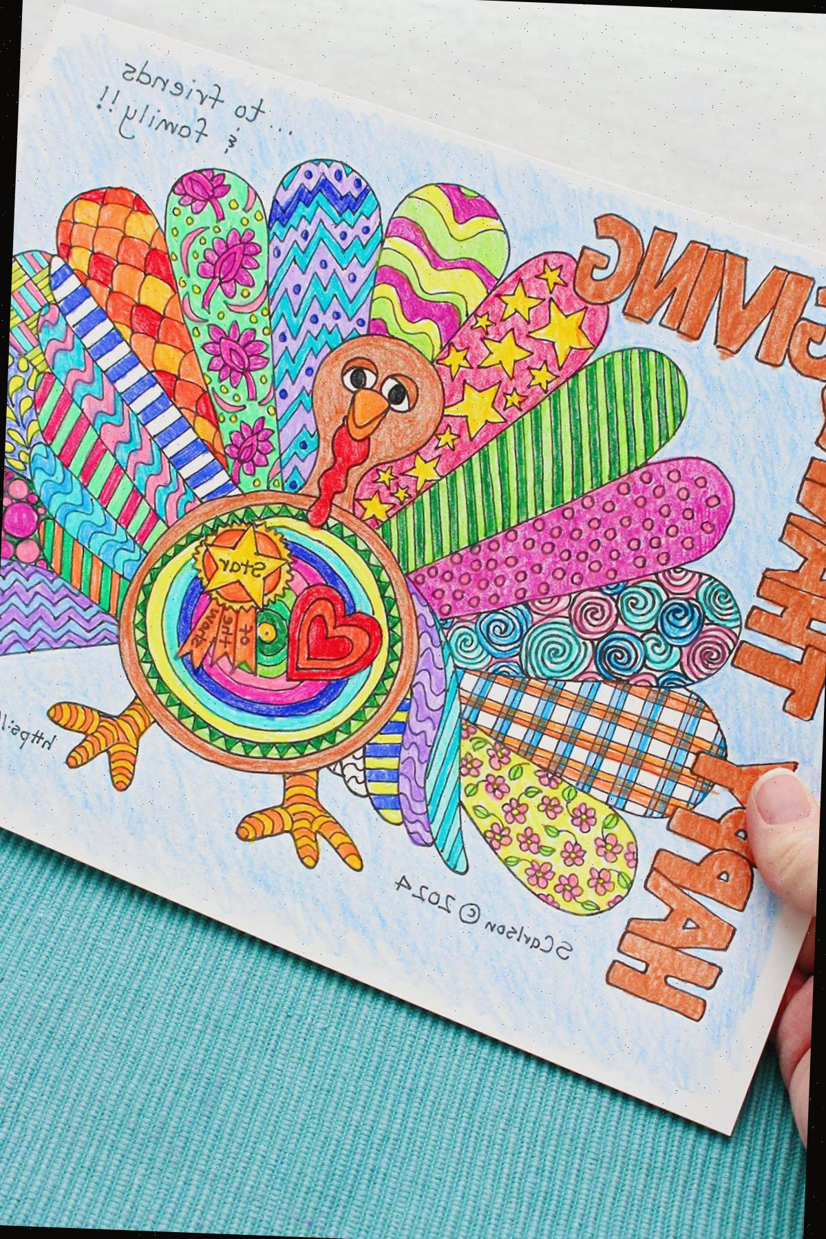 Uma pessoa segurando uma página de colorir de Ação de Graças exibindo 'Feliz Ação de Graças' e um peru com asas decoradas e coloridas.