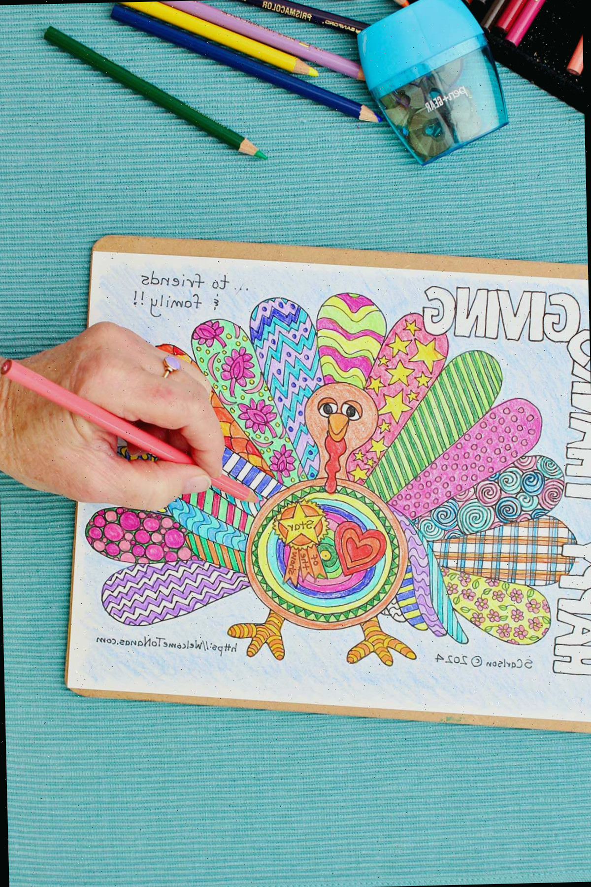 Página de colorir de Ação de Graças de um peru com asas decoradas, fixada a uma prancheta sobre um lugar-manto aqua, acompanhada de lápis de cor. Pessoa finalizando os detalhes nas penas do peru.
