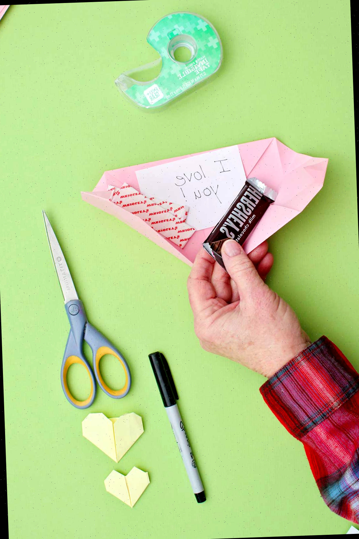 Uma pessoa colocando uma pequena barra de chocolate e um bilhete dentro de um coração de origami rosa.