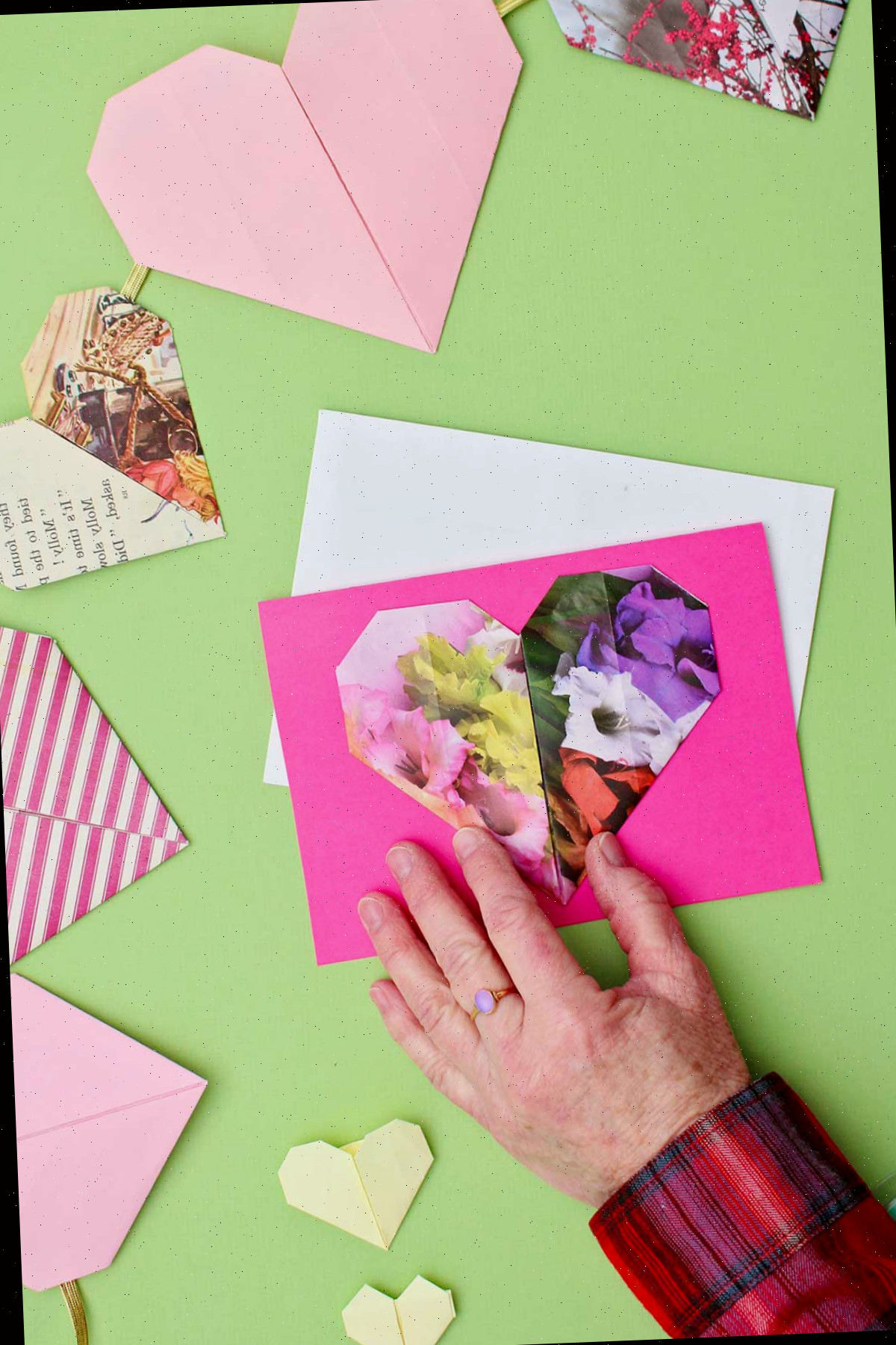 Um coração de origami floral sendo posicionado em um cartão rosa com outros corações de origami nas proximidades.