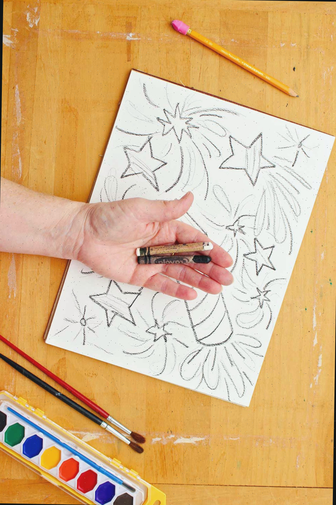 Mão segurando um lápis de cera preto e branco acima de um desenho de estrelas e foguetes.