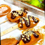 Fatias de maçã caramelada finalizadas no palito descansando sobre papel manteiga. Algumas têm espirais de chocolate amargo e nozes salpicadas.